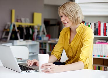 Woman in yellow shirt working on laptop at desk with bookshelf behind her