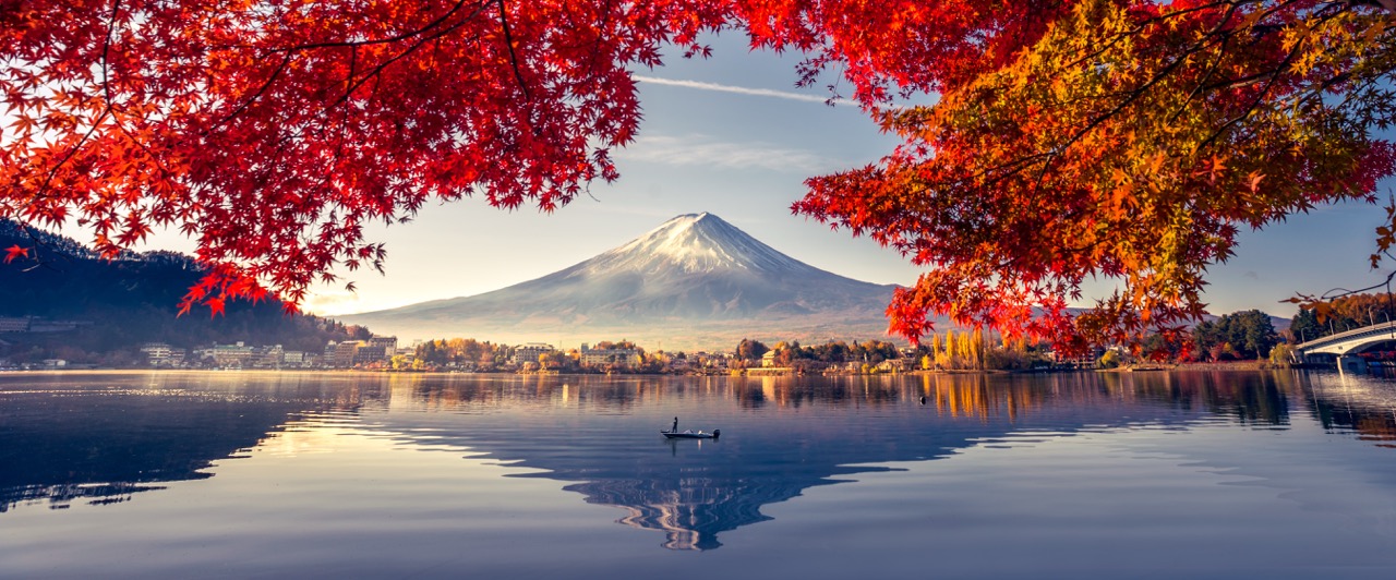Mount Fuji snow-capped peak reflected in calm lake framed by vibrant red autumn maple leaves