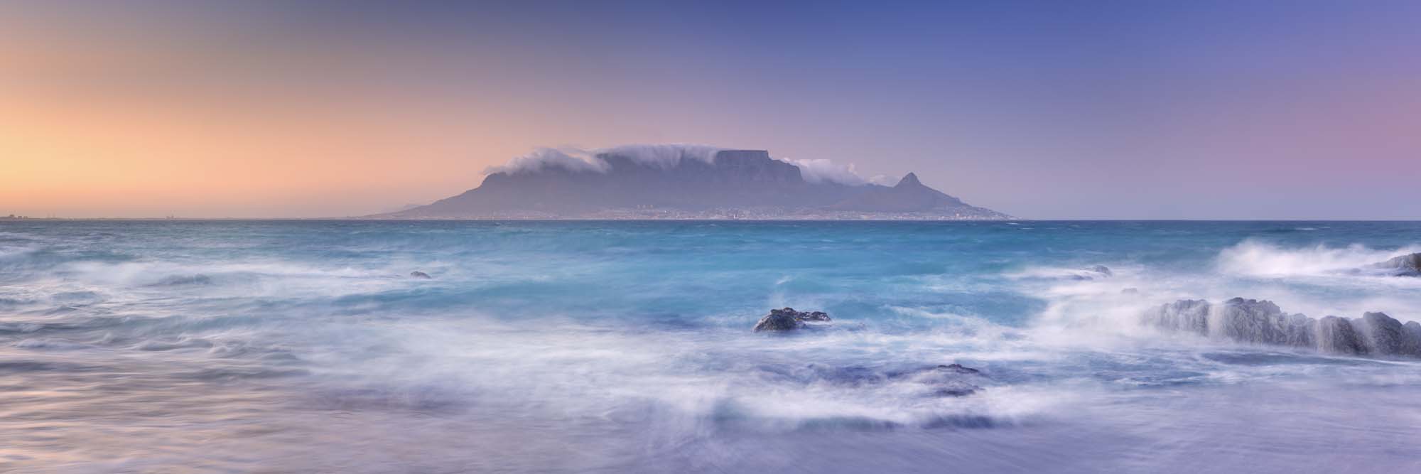 Table Mountain at sunrise across ocean with rolling waves and rocky shore