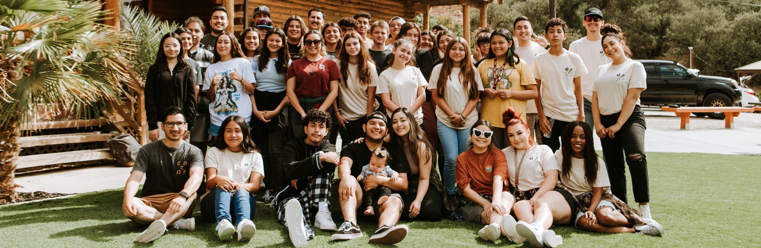 Large diverse group of young adults posing together on grass outside wooden lodge building