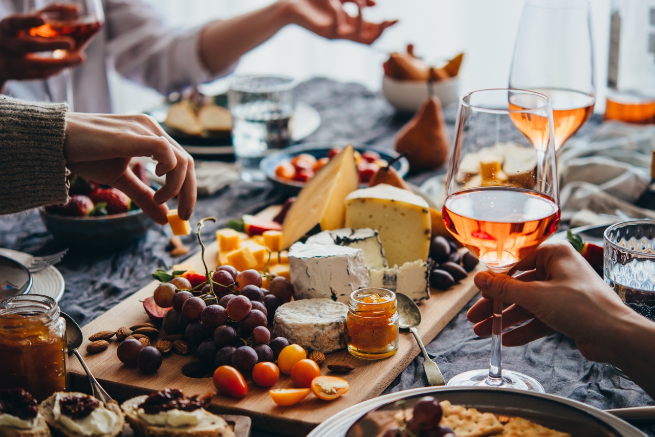 Hands reaching for cheese and grapes at a wooden board with wine glasses and charcuterie spread