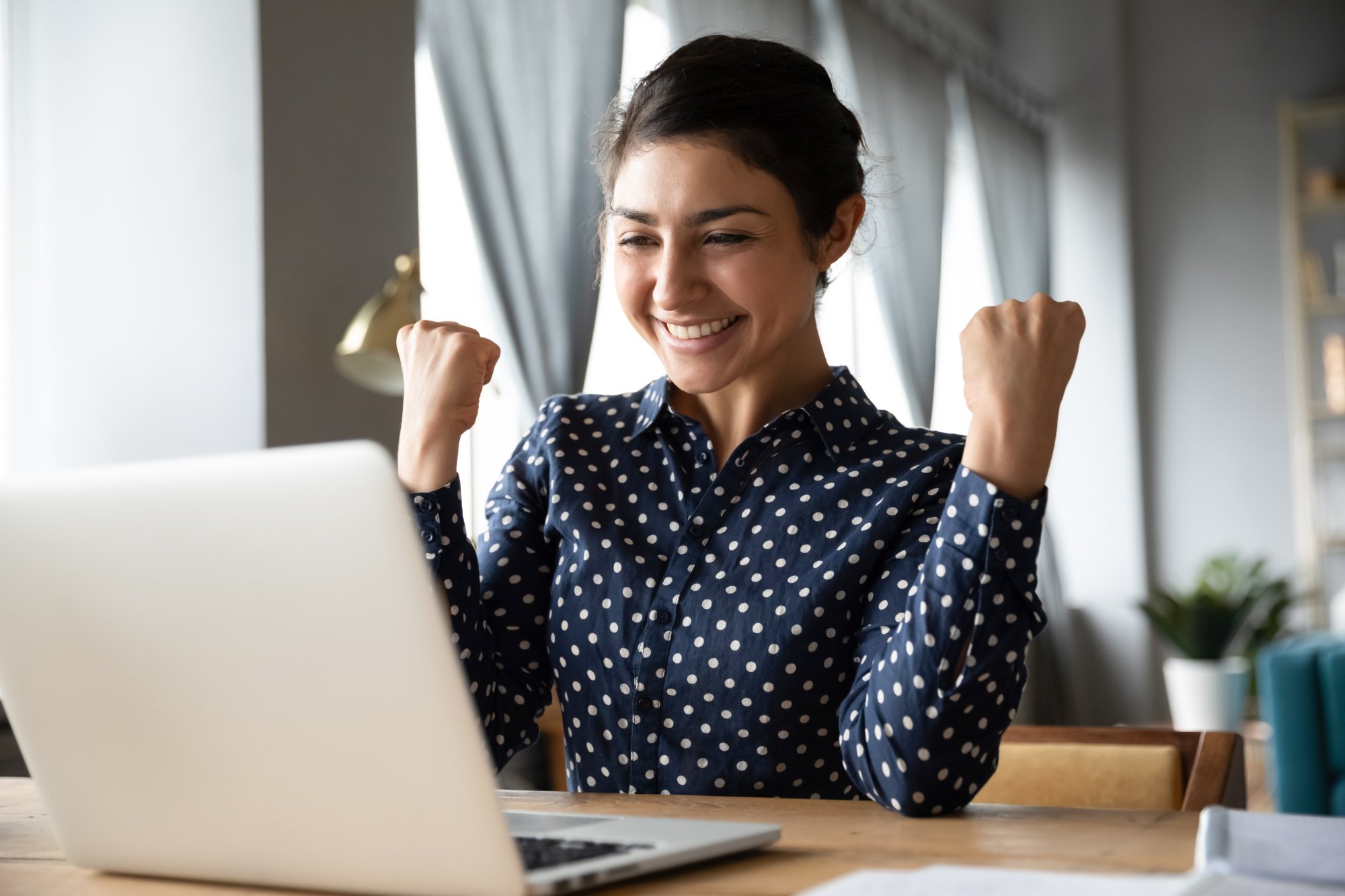A smiling young woman in a navy polka-dot blouse sits at a wooden desk with a laptop, raising both